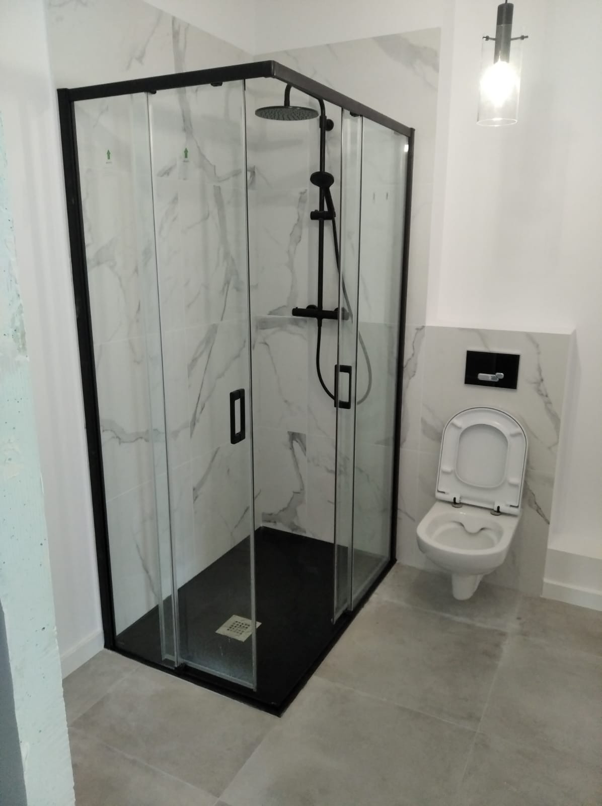 Nowoczesna kabina prysznicowa z czarnymi profilami i deszczownicą, wykończona płytkami imitującymi marmur, obok toaleta wisząca.
