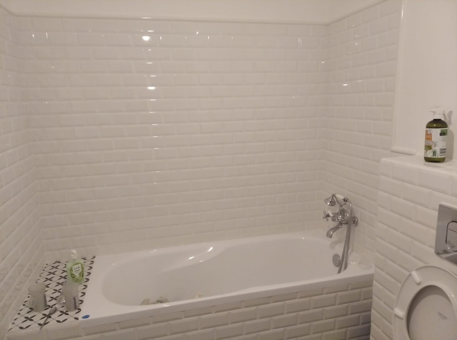 Biała wanna obudowana płytkami typu metro, z chromowaną baterią i prysznicem, otoczona ścianami wyłożonymi tymi samymi płytkami, z widocznym fragmentem toalety i butelką płynu.