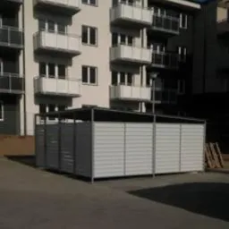 Metalowa konstrukcja osłaniająca pojemniki na śmieci, panelowa zabudowa z poziomych listew, na tle nowego bloku mieszkalnego z białymi balkonami.