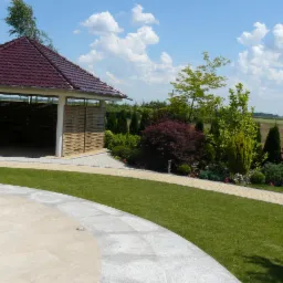 Zadbany ogród z altaną krytą czerwoną dachówką, równo przystrzyżony trawnik, ścieżki z kostki brukowej i różnorodne drzewa oraz krzewy ozdobne.