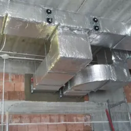 System kanałów wentylacyjnych w izolacji aluminiowej, zamontowany pod betonowym stropem, widoczne przewody elektryczne i fragment ściany z cegły.