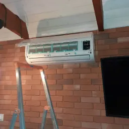 Montaż klimatyzacji w pomieszczeniu z ceglaną ścianą i drewnianym sufitem, widoczna drabina aluminiowa podczas instalacji.