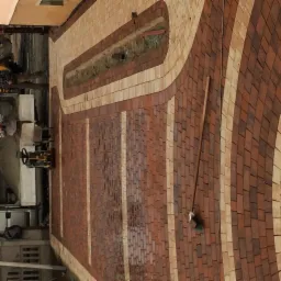 Wzór z czerwono-beżowej nawierzchni ułożonej z drobnych elementów, widoczna ubijarka mechaniczna i drewniany kij z zieloną końcówką.