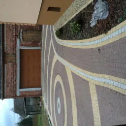 Ułożona kostka brukowa w odcieniach brązu i beżu tworząca wzór koncentrycznych okręgów na podjeździe, obok ceglany budynek z drewnianą bramą garażową.
