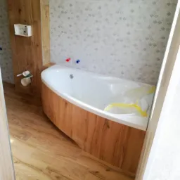 Nowoczesna, narożna wanna obudowana drewnopodobną płytą, z widocznymi przyłączami wody na tle jasnoszarej mozaiki ściennej, na podłodze imitacja drewna, częściowo zabezpieczona folią ochronną.