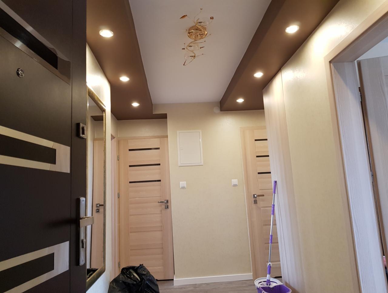 Korytarz po remoncie: podwieszany sufit z brązowymi wnękami i punktowym oświetleniem, widoczne drzwi wewnętrzne z poziomymi wstawkami, mop i wiadro po sprzątaniu.