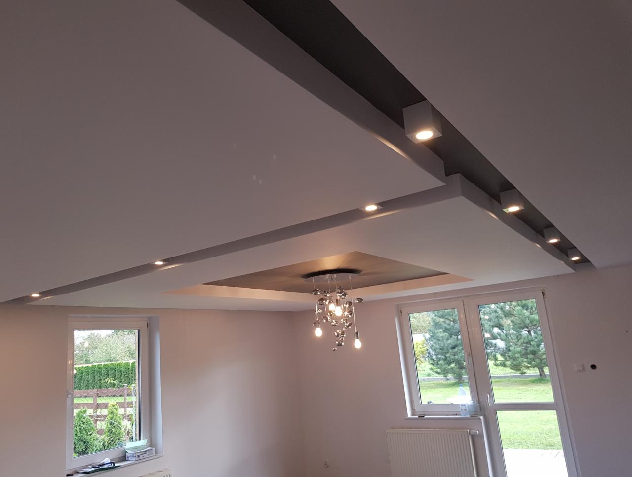 Sufit podwieszany z oświetleniem punktowym i centralnym żyrandolem, widok na okna z ogrodem w tle.