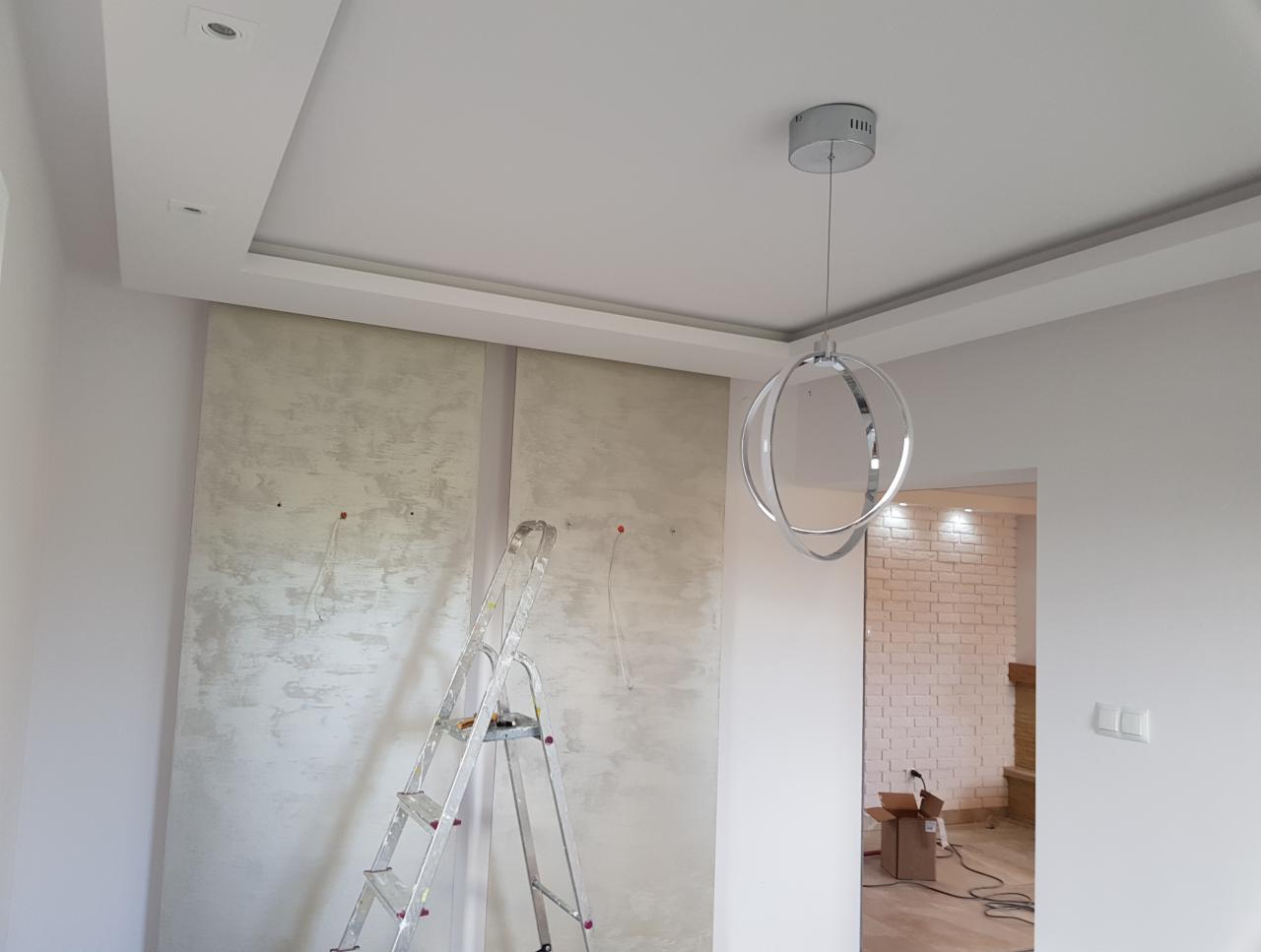 Ściana z fakturą w trakcie wykańczania, widoczna drabina aluminiowa, minimalistyczna lampa wisząca i fragment ceglanej ściany w tle.