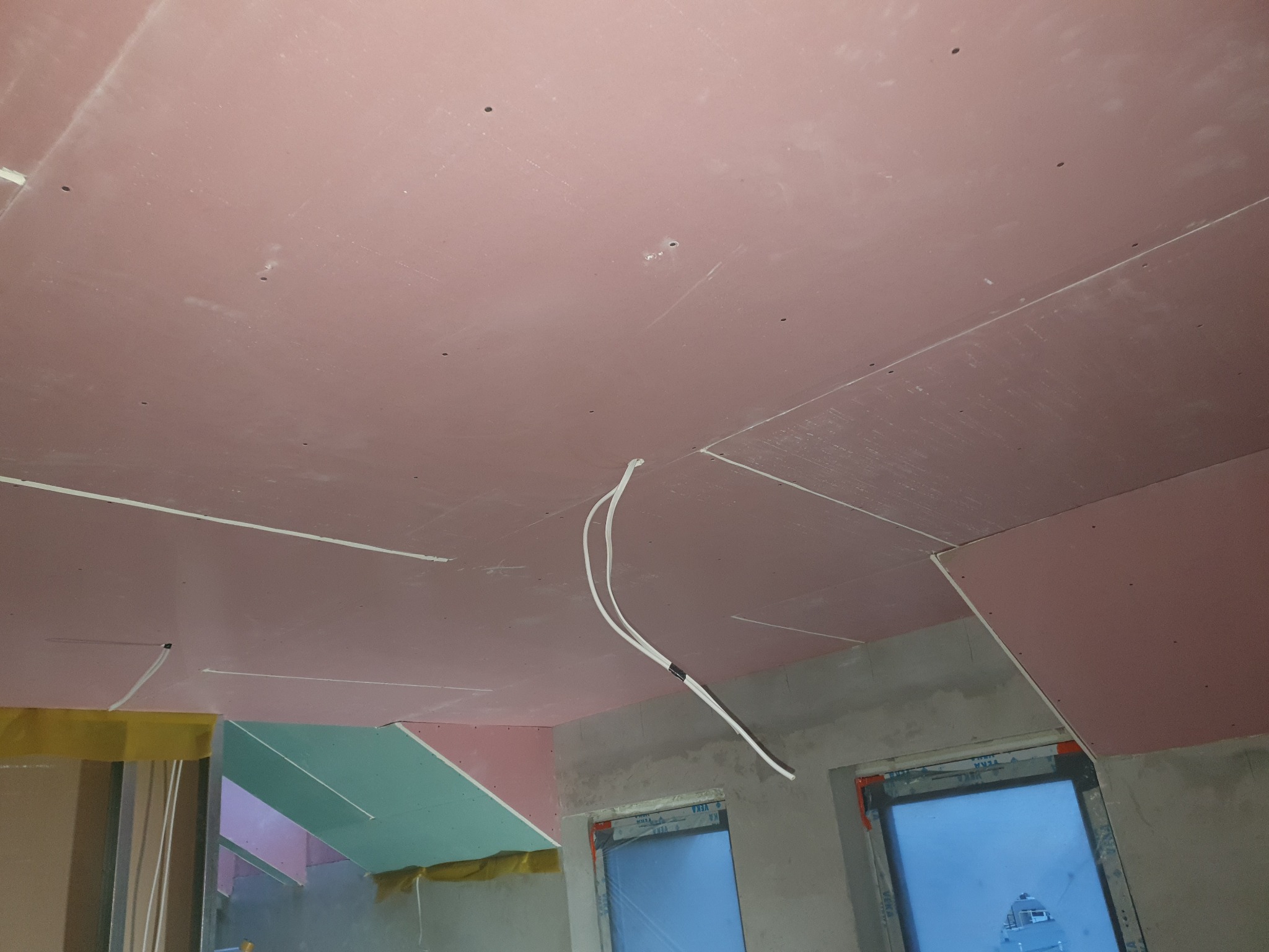 Montaż sufitu podwieszanego z płyt w kolorze różowym, widoczne kable elektryczne, okna w stanie surowym, taśma maskująca na łączeniach płyt.