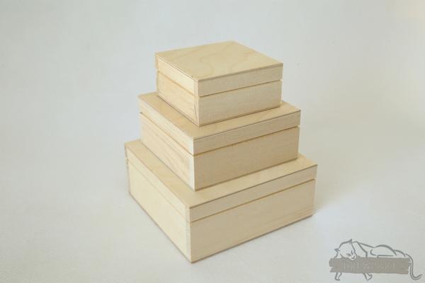 Trzy drewniane, surowe pudełka różnej wielkości ułożone jedno na drugim, od największego u dołu do najmniejszego u góry, na jasnym tle.