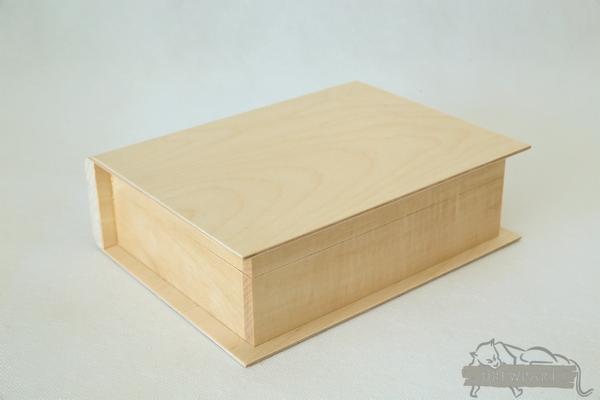 Prostokątne, jasne drewniane pudełko z pokrywką, przypominające książkę, na jasnym tle.