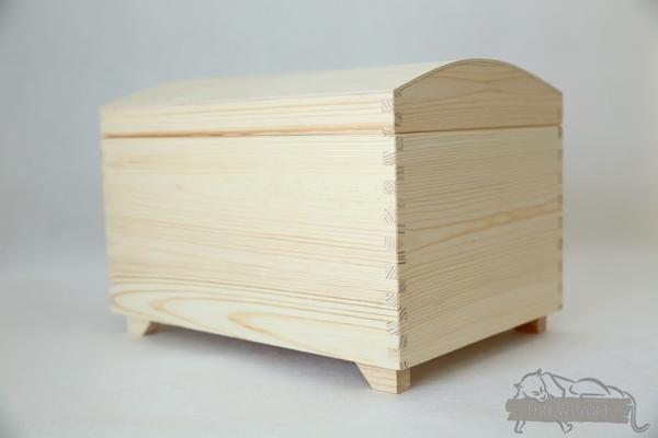 Drewniana skrzynia z jasnego surowego drewna, widoczne połączenia na wczepy proste, zaokrąglone wieko i małe nóżki.