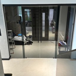 TNS-Glass - Szklane drzwi przesuwne z czarnymi ramami w nowoczesnym wnętrzu. Widoczne odbicie pomieszczenia z drabiną i narzędziami.