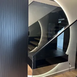 Nowoczesna balustrada szklana z podświetleniem LED, widoczna z perspektywy schodów. Czarna ściana z lamelami po lewej stronie kadru.