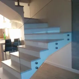 Schody drewniane z białymi podstopnicami i szklaną balustradą mocowaną punktowo, widoczne elementy kuchni w tle.