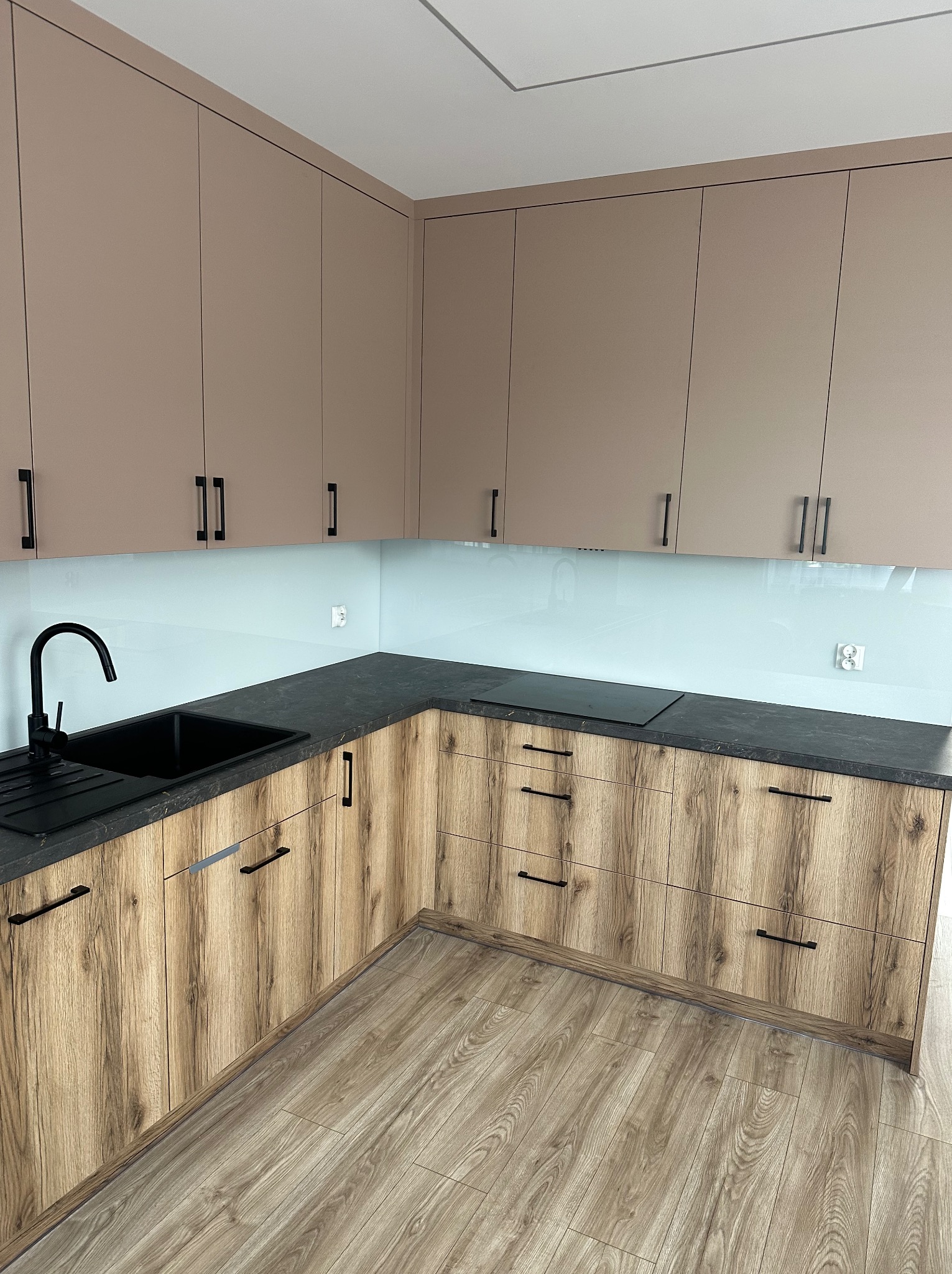 Nowoczesna kuchnia z drewnianymi szafkami, czarnym blatem i jasnoniebieskim panelem szklanym nad blatem. Minimalistyczne uchwyty i armatura w czarnym kolorze.