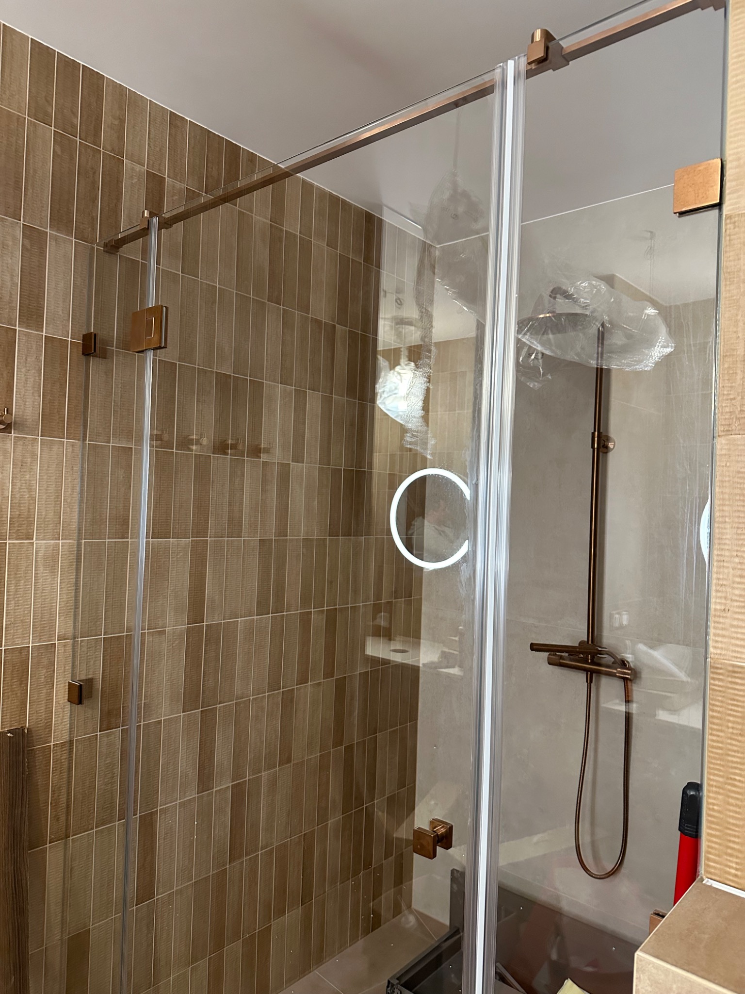 Szklana kabina prysznicowa z brązowymi okuciami na tle beżowych płytek. Widoczne elementy montażowe i słuchawka prysznicowa w folii ochronnej.