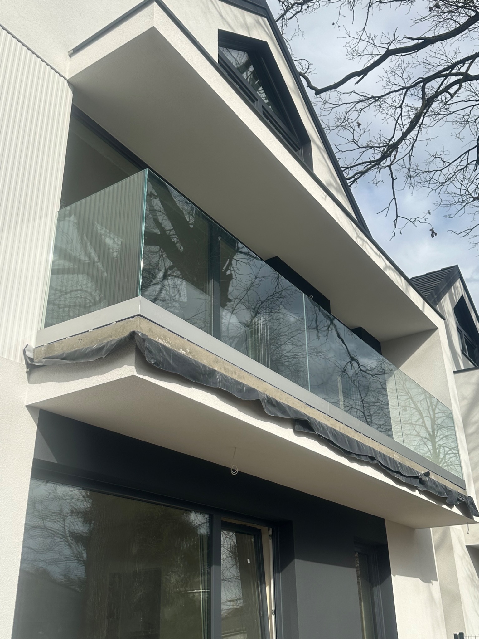 Nowoczesna balustrada ze szkła na balkonie budynku z białą elewacją. Widoczne detale montażowe i odbicie drzew w szkle. Minimalistyczny design.