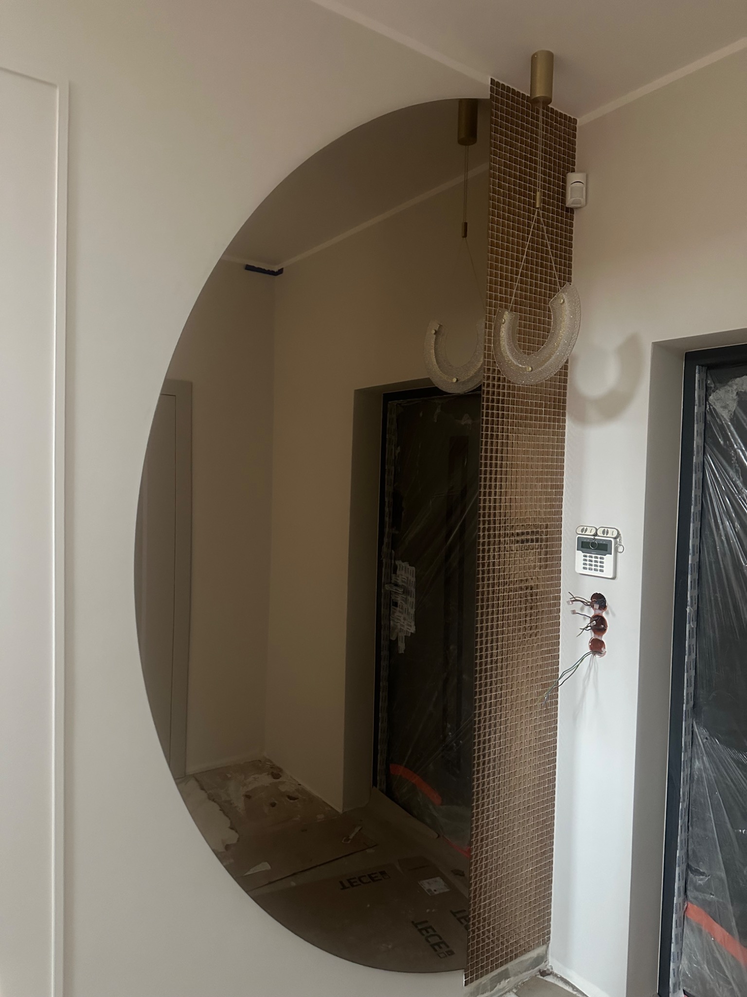 Okrągłe lustro wkomponowane w ścianę, odbijające wnętrze. Obok mozaika z brązowych płytek i wiszące lampy. Widoczne elementy wykończenia wnętrz w trakcie prac.