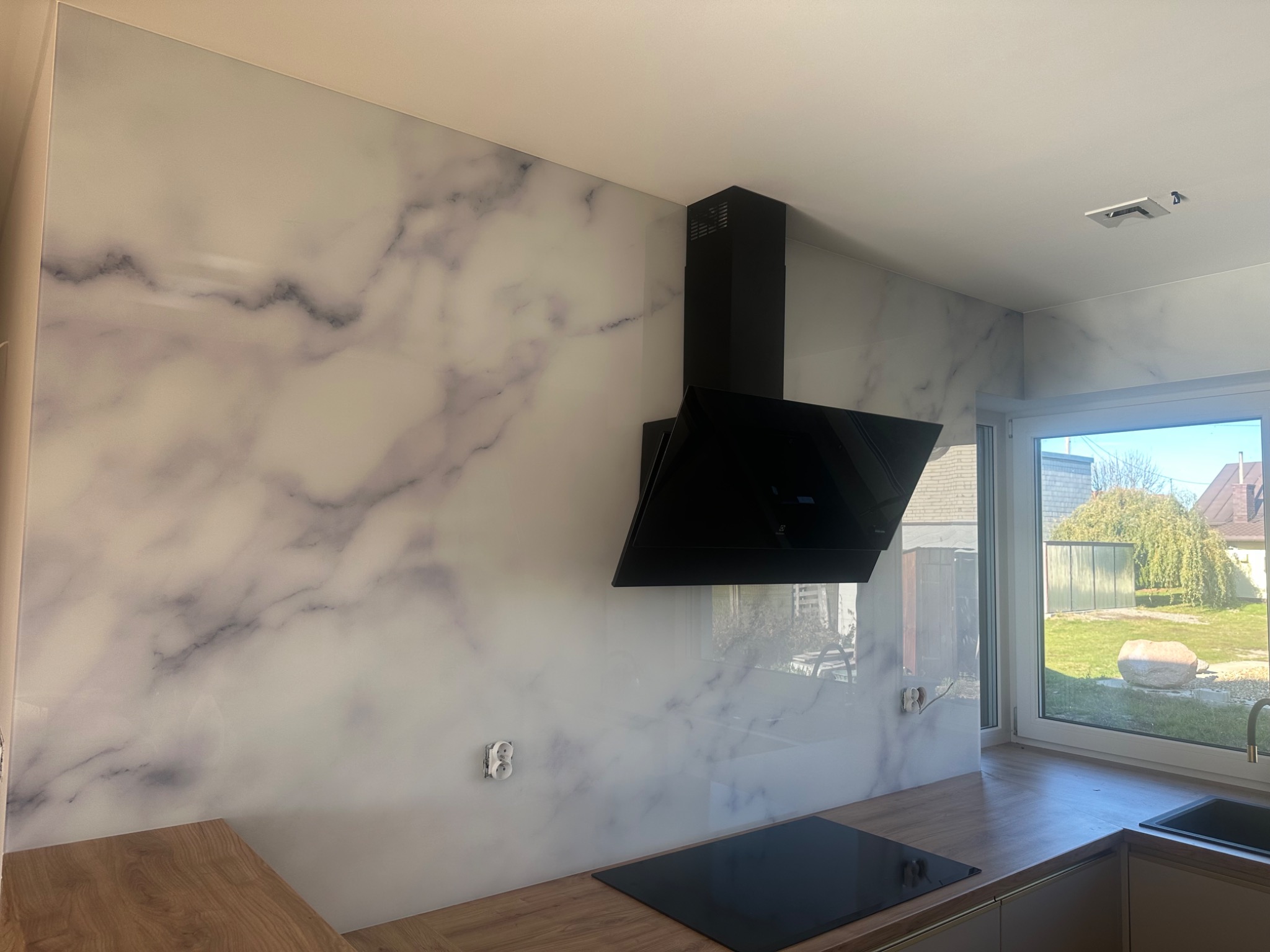 Szklana ściana w kuchni z wzorem marmuru, czarny okap, drewniany blat, widok przez okno na zewnątrz. Nowoczesne wykończenie wnętrza, gniazdko elektryczne na ścianie.