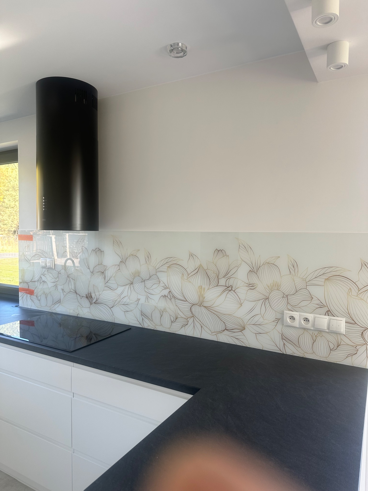 Kuchenny panel szklany z motywem kwiatowym, czarny okap cylindryczny, białe szafki i czarny blat. Nowoczesna kuchnia z elementami dekoracyjnymi.