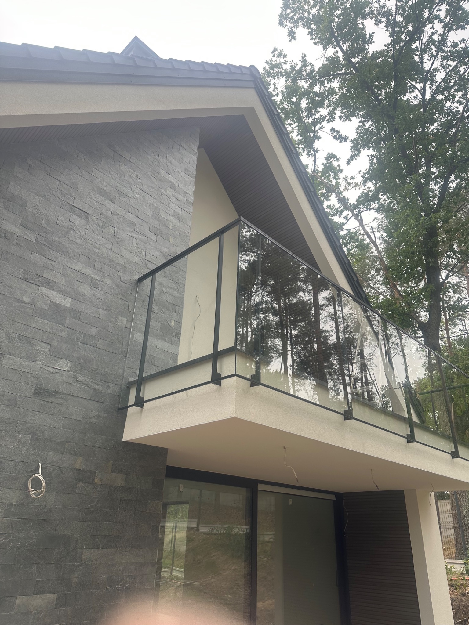 Nowoczesny balkon z czarną ramą i szklaną balustradą, widok z dołu, na tle ściany z szarej cegły i zieleni drzew. Minimalistyczny design, naturalne światło.