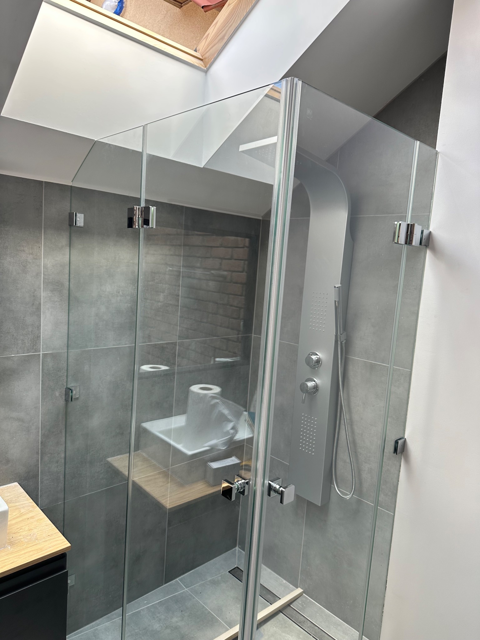 Szklana kabina prysznicowa z deszczownicą, montowana na zawiasach, w nowoczesnej łazience z szarą glazurą i oknem dachowym.