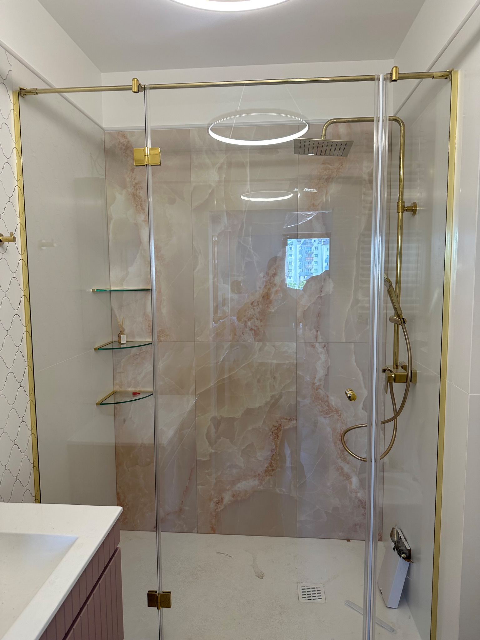Nowoczesna kabina prysznicowa z transparentnego szkła, złote okucia, marmurowe płytki w odcieniach beżu i różu, szklane półki. Eleganckie wykończenie łazienki.