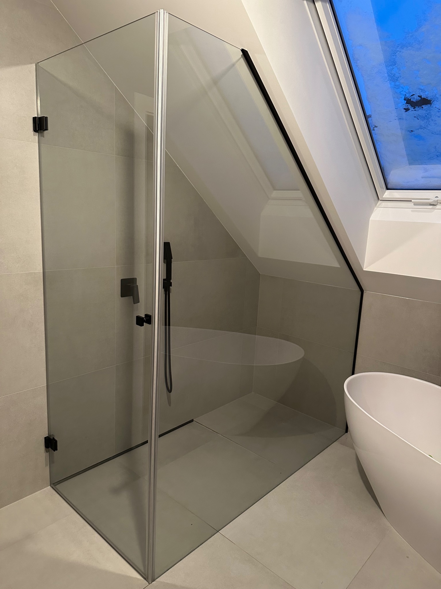 Szklana kabina prysznicowa z czarną armaturą w nowoczesnej łazience na poddaszu z oknem. Minimalistyczny design, jasne płytki, wanna wolnostojąca.