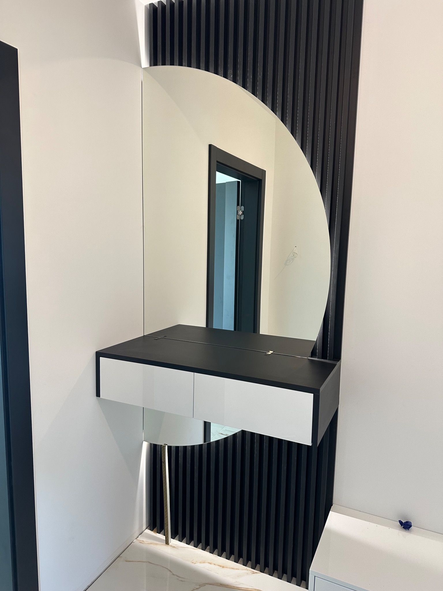 Półokrągłe lustro z czarną konsolą i białymi szufladami, wsparte na złotej nóżce. Dekoracyjne czarne listwy na ścianie. Widok na wnętrze mieszkania w odbiciu.