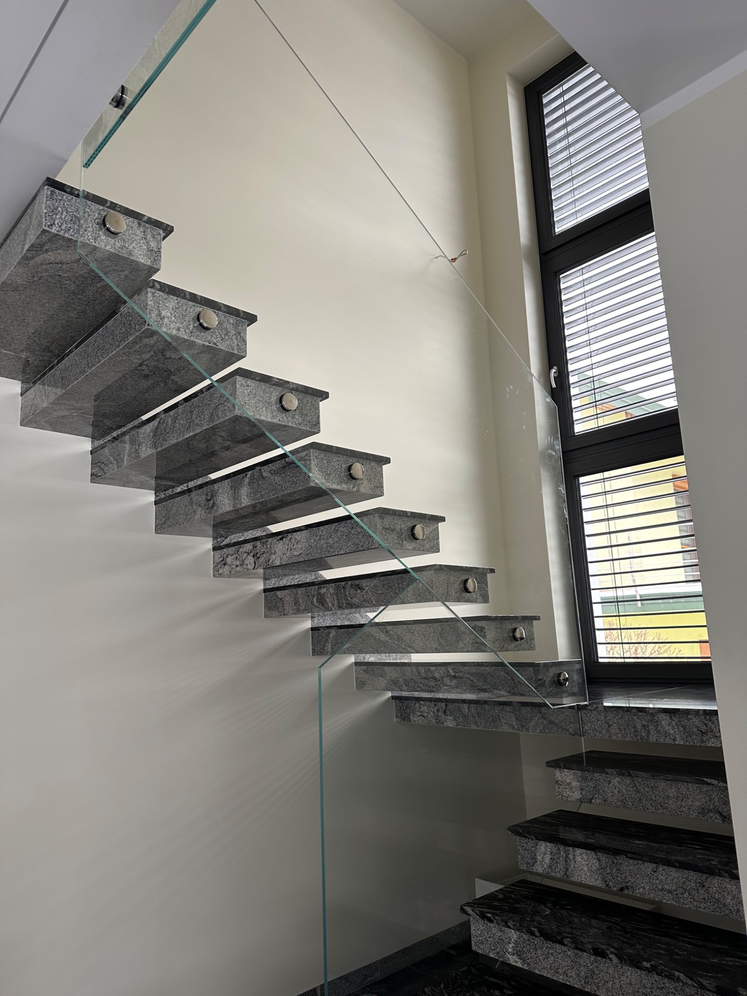 Nowoczesne schody z granitu ze szklaną balustradą, mocowaną na metalowych uchwytach. Widok od dołu, z oknem w tle.