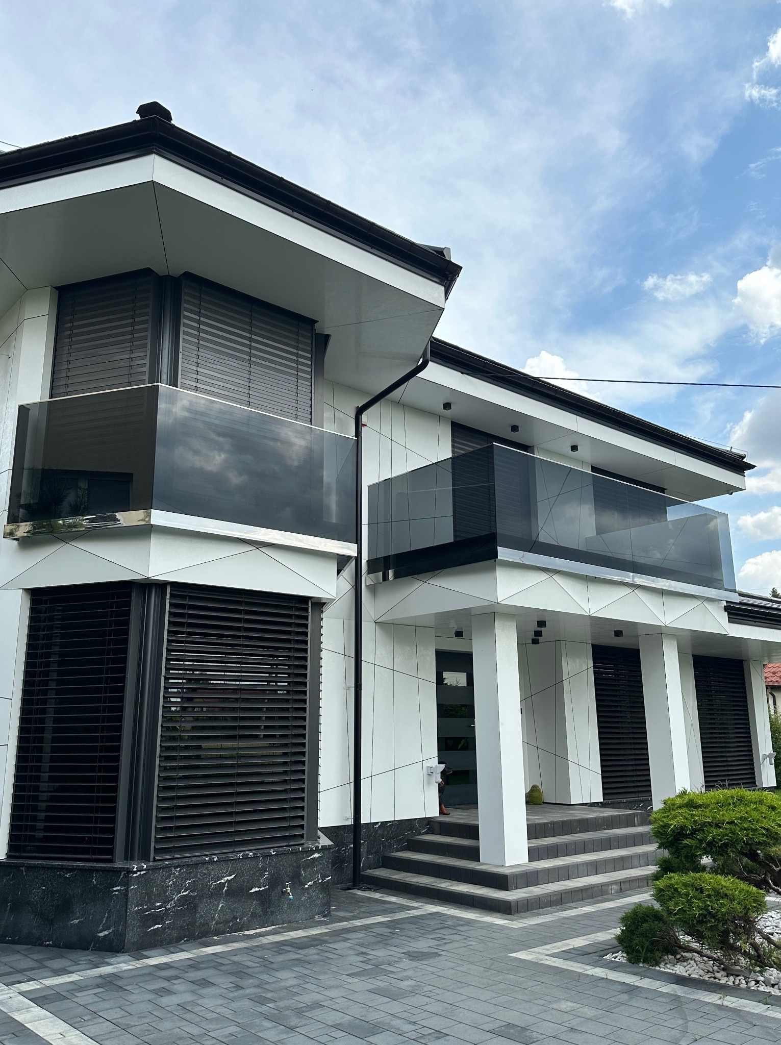 Nowoczesny dom z białymi panelami i czarnymi balustradami szklanymi na balkonach, widok z dołu w słoneczny dzień. Eleganckie wykończenie i geometryczny design.