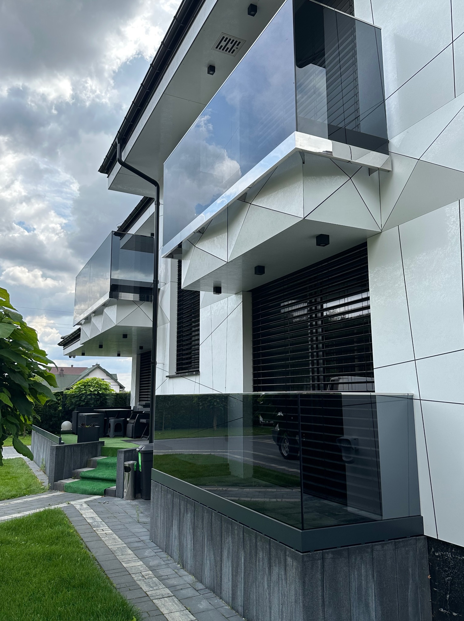 Nowoczesny dom z balkonami i balustradami ze szkła, widok z dołu. Elewacja w geometryczne wzory, szare balustrady, odbicie nieba w szkle.