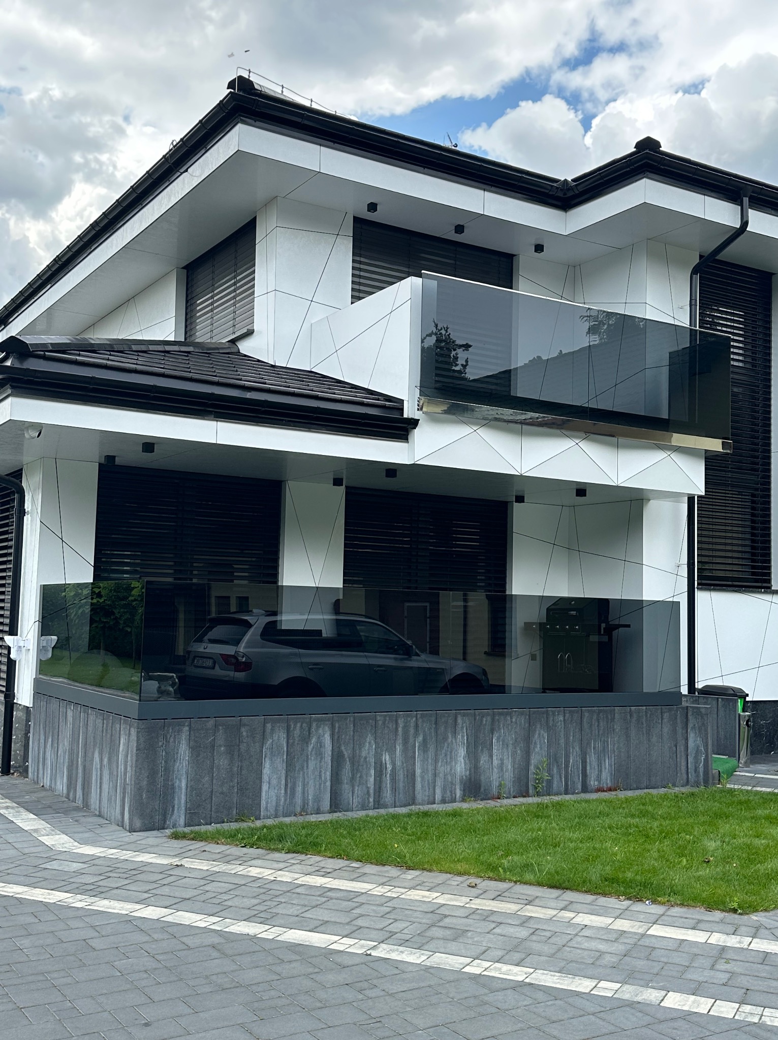 Nowoczesny dom z szklaną balustradą balkonową i tarasową. Widoczny fragment elewacji, okna z żaluzjami i samochód za szkłem. Minimalistyczny design.