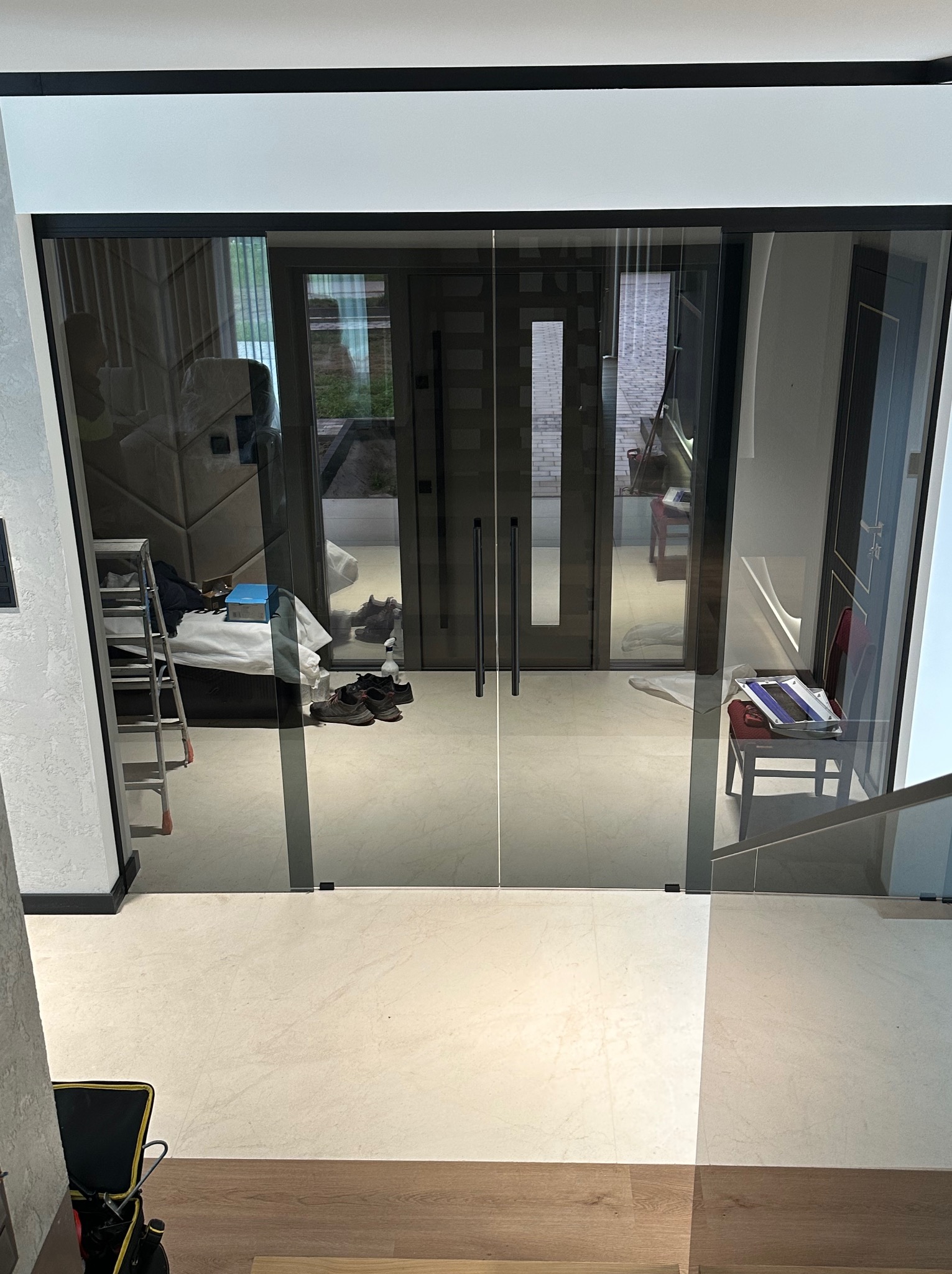 Szklane drzwi przesuwne z czarnymi ramami w nowoczesnym wnętrzu. Widoczne odbicie pomieszczenia z drabiną i narzędziami.