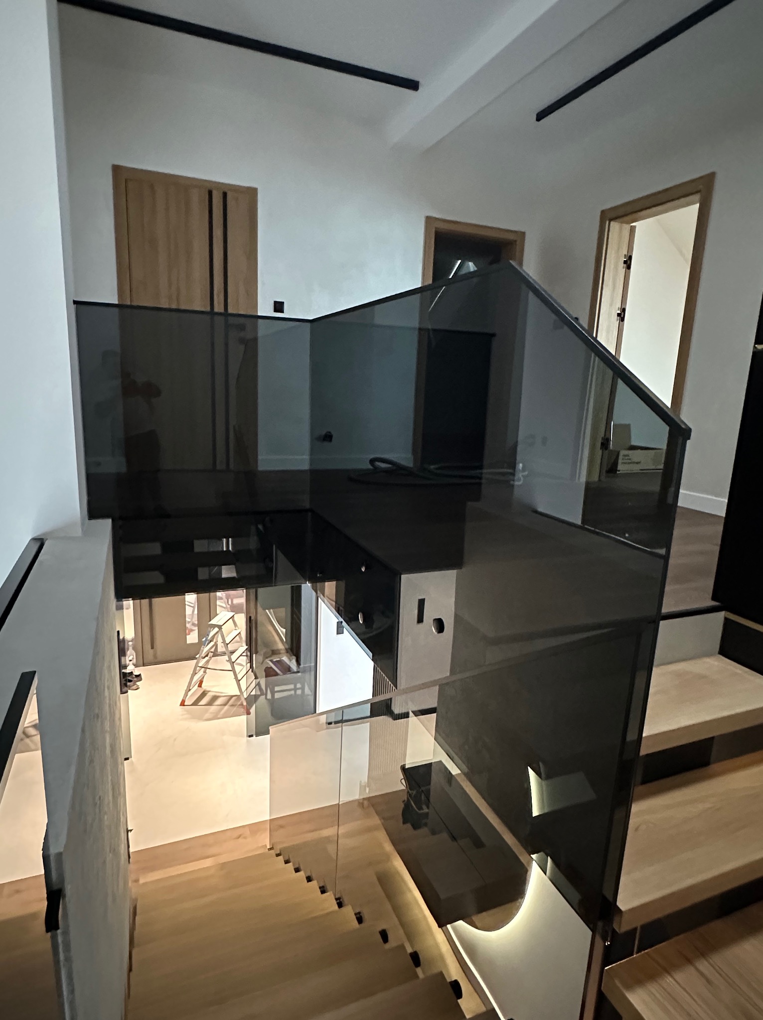 Nowoczesna, grafitowa balustrada szklana na drewnianych schodach w jasnym wnętrzu. Widoczne drzwi i drabina malarska w tle. Minimalistyczny design.