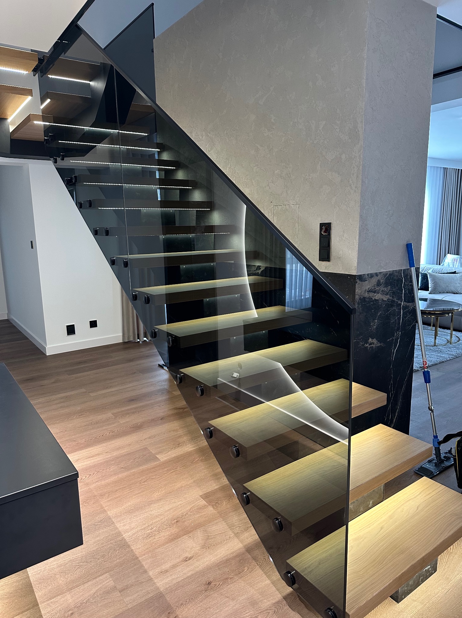 Nowoczesne schody z drewnianymi stopniami i szklaną balustradą, podświetlane diodami LED. Czarny marmur na filarze, minimalistyczny design.