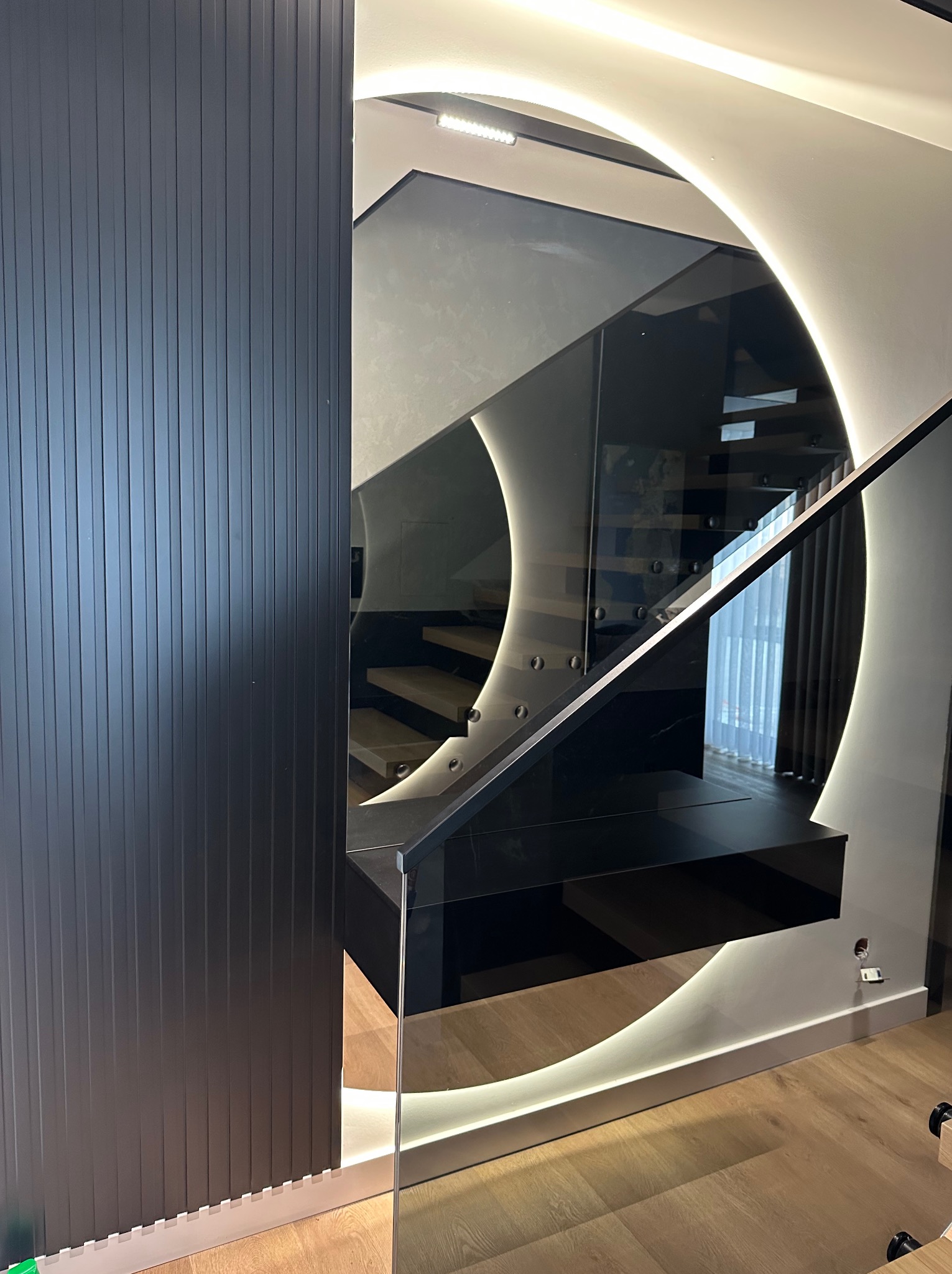 Nowoczesna balustrada szklana z podświetleniem LED, widoczna z perspektywy schodów. Czarna ściana z lamelami po lewej stronie kadru.