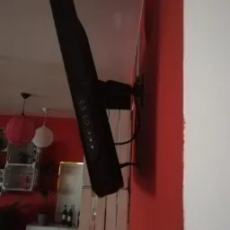 Czarny panel sterowania domofonu/wideofonu zamontowany na czerwonej ścianie, widoczne kable i dekoracyjne elementy wnętrza w tle.