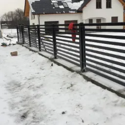 Czarny, poziomy ogrodzenie ze stalowych profili w trakcie montażu, widoczny samochód z otwartym bagażnikiem i narzędzia, śnieg na ziemi.