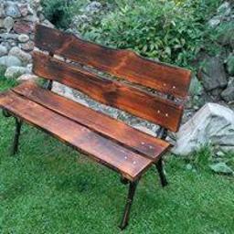 ArtGard - Rustykalna ławka ogrodowa z ciemnego drewna o nieregularnych krawędziach, osadzona na metalowych nogach, na tle zielonej trawy i kamiennego murku.