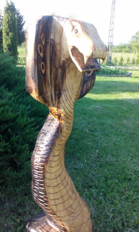 Drewniana rzeźba przedstawiająca kobrę w ogrodzie, widoczne detale wykonania i naturalne otoczenie.