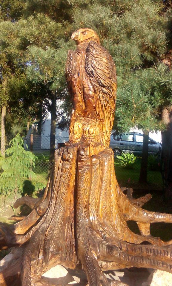 Drewniana rzeźba orła stojąca na pniu drzewa z widocznymi korzeniami, umieszczona w ogrodzie na tle drzew iglastych i ogrodzenia.