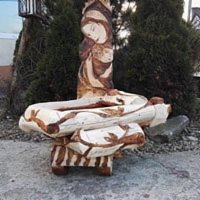 Ręcznie rzeźbiony, drewniany kwietnik ogrodowy z motywem roślinnym, umieszczony na betonowym podłożu, na tle iglaków.