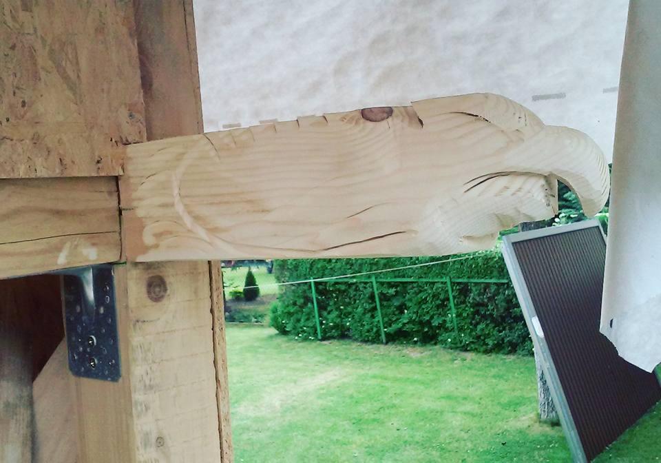 Surowe drewno rzeźbione w kształt głowy orła, przymocowane do drewnianej konstrukcji na zewnątrz, z widokiem na trawnik i zielony żywopłot w tle.