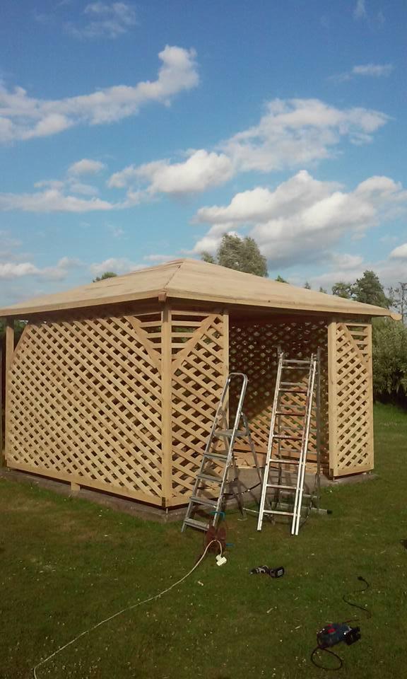 Drewniana altana ogrodowa w trakcie budowy, z widocznymi drabinkami i narzędziami na trawie, na tle błękitnego nieba z chmurami.