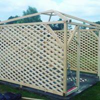 Surowa drewniana konstrukcja altany z kratownicowymi ścianami w Bogumiłowicach, widoczna drabina wewnątrz. Konstrukcja w trakcie budowy, bez dachu.