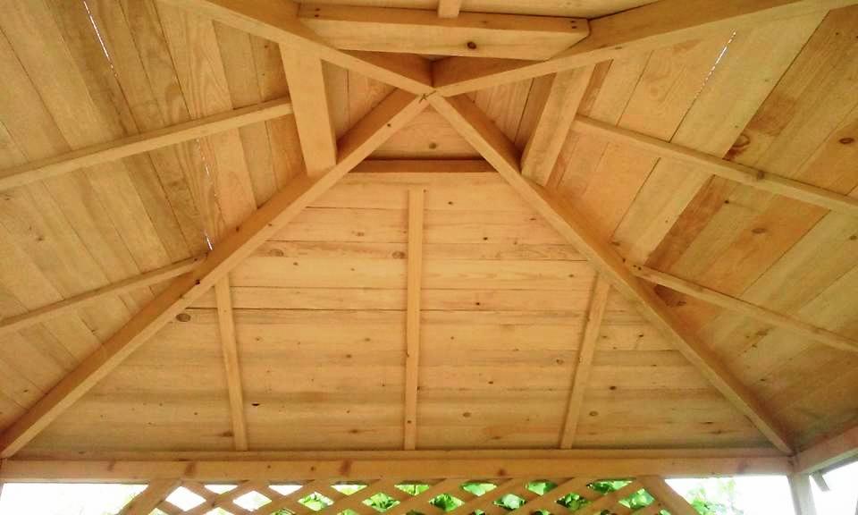 Drewniana konstrukcja dachu altany ogrodowej z widocznymi belkami i deskami, z fragmentem ażurowej ścianki w tle.