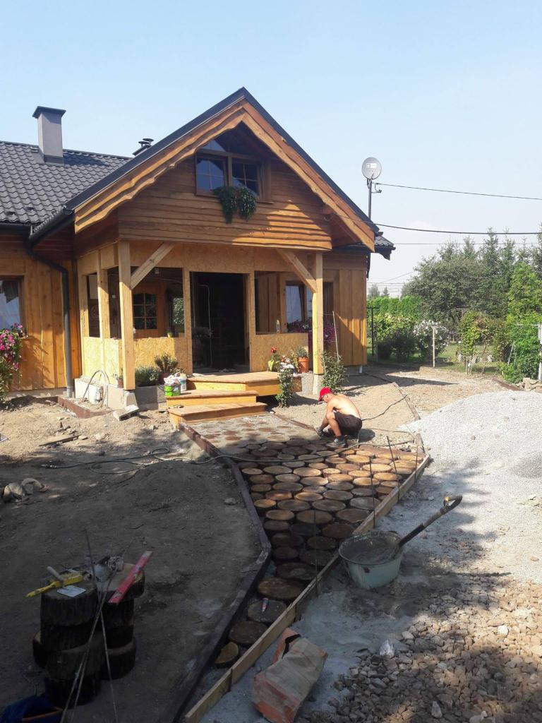 Układanie ścieżki z drewnianych krążków prowadzącej do drewnianego domu z werandą, widoczny mężczyzna w czerwonej czapce podczas pracy, narzędzia i materiały budowlane w tle.