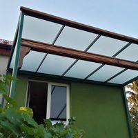 Zadaszenie nad oknem z brązową drewnianą ramą i przezroczystymi panelami, wsparte na zielonych słupach, widoczne z dołu, z zielonym krzewem w rogu kadru.